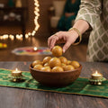 Woman handpicking a besan laddu from a bowl set against a festive decoration