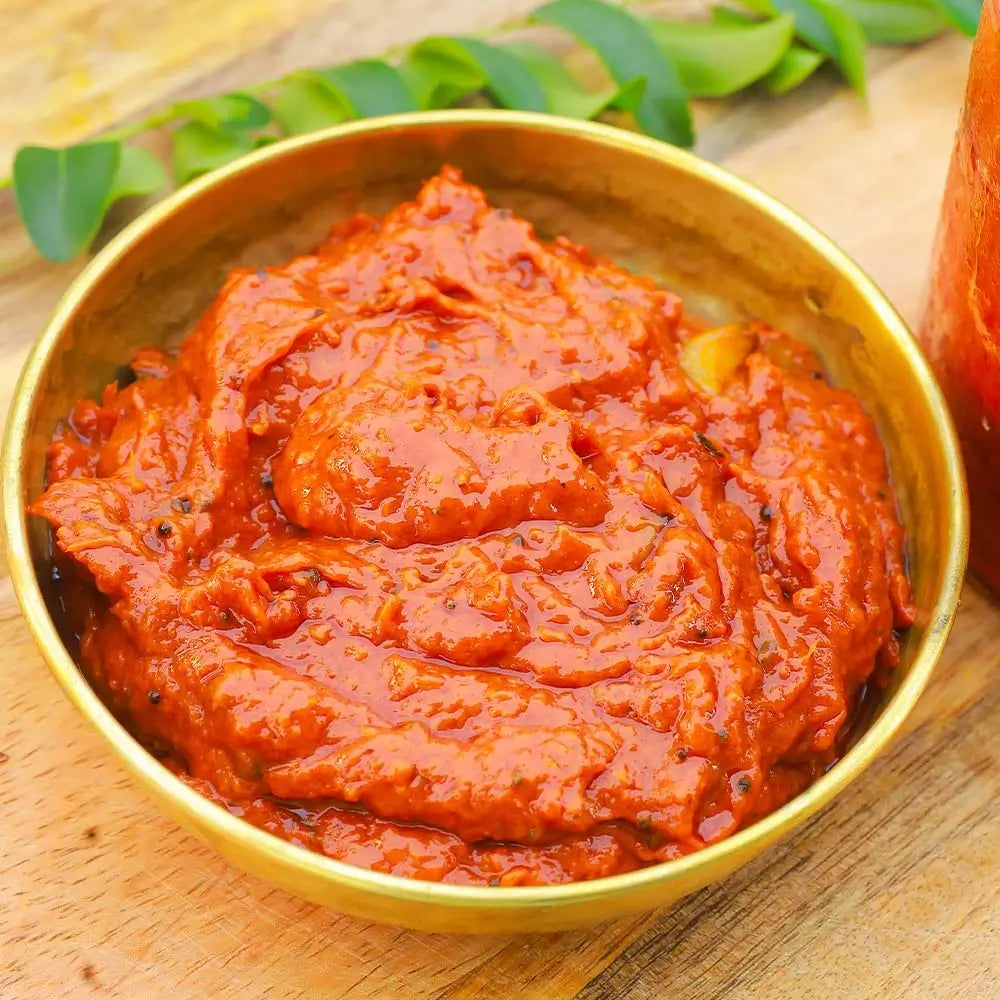 Red tomato pickle in a gold bowl on a wooden surface with green leaves in the background