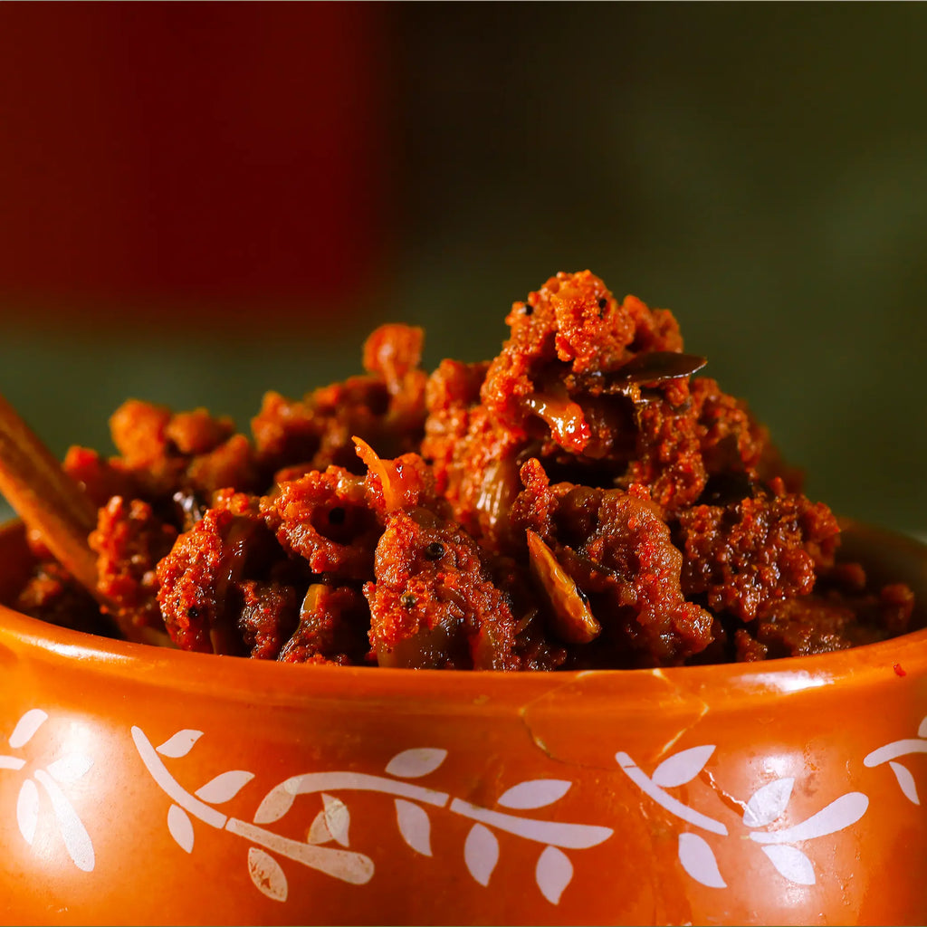 A close-up shot of a textured, reddish-brown cauliflower pickle served in a terracotta-coloured bowl with white leaf patterns, set against a dark, blurred background.