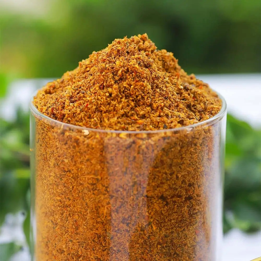 Close-up of a glass container filled with brown avise ginjala podi spice mix on a blurred green background