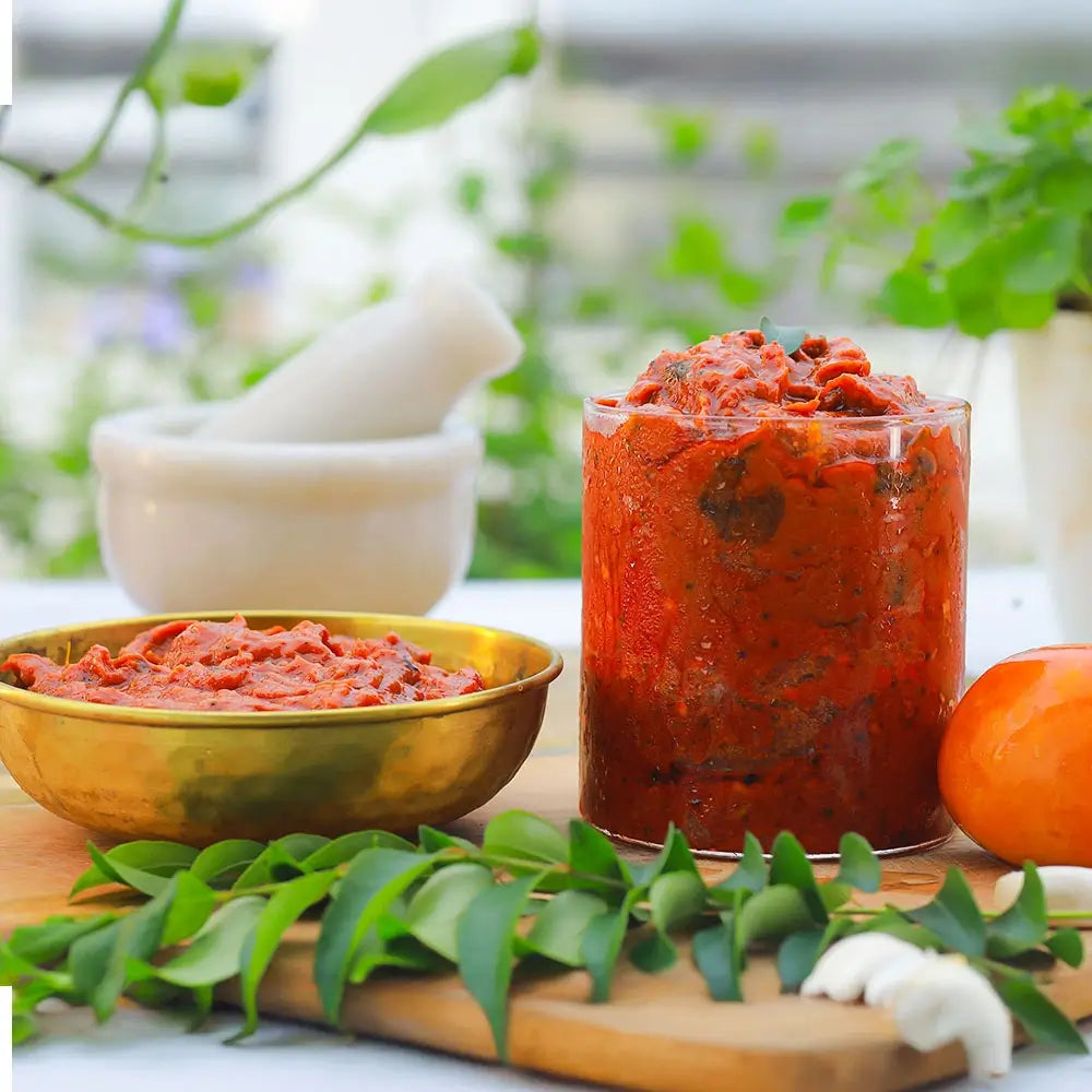 Red tomato pickle in a glass jar and bowl with green leaves and an orange on a wooden board.