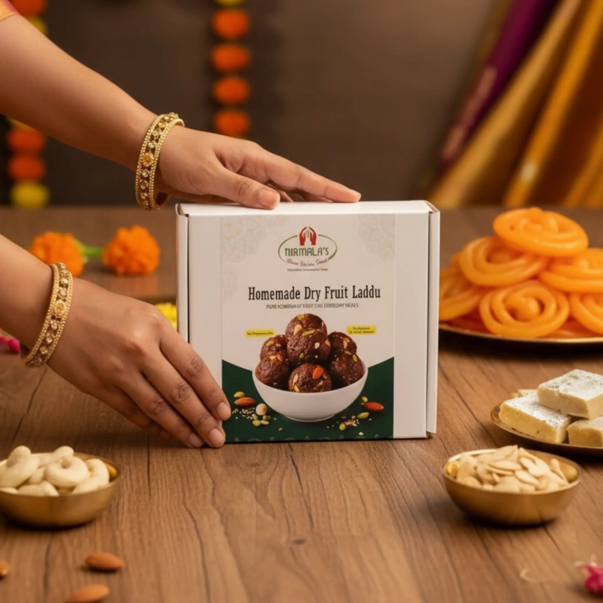 Box of 'Homemade Dry Fruit Laddu' with hands reaching towards it on a wooden table with snacks.