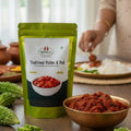 Green package of 'Nirmala's Traditional Pickles & Podi' on a table with a bowl of kakarakaya pickle and green leaves.