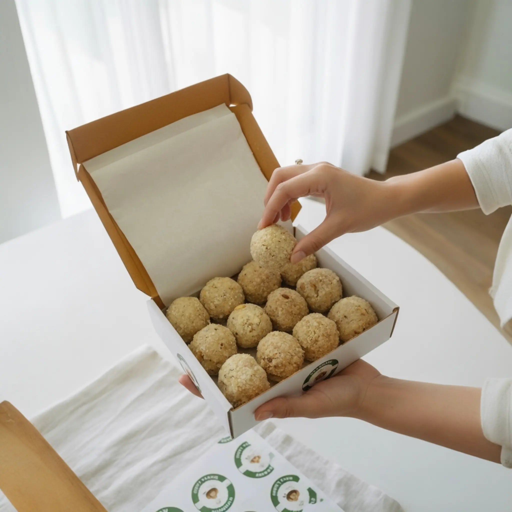 Person holding a box of round, brown food items on a white surface with a light curtain background.