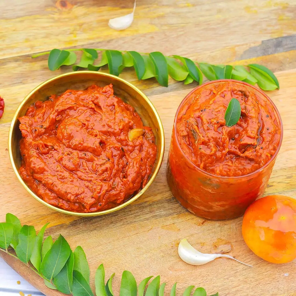 Red tomato pickle in a gold bowl and glass container on a wooden surface with green leaves and a tomato.