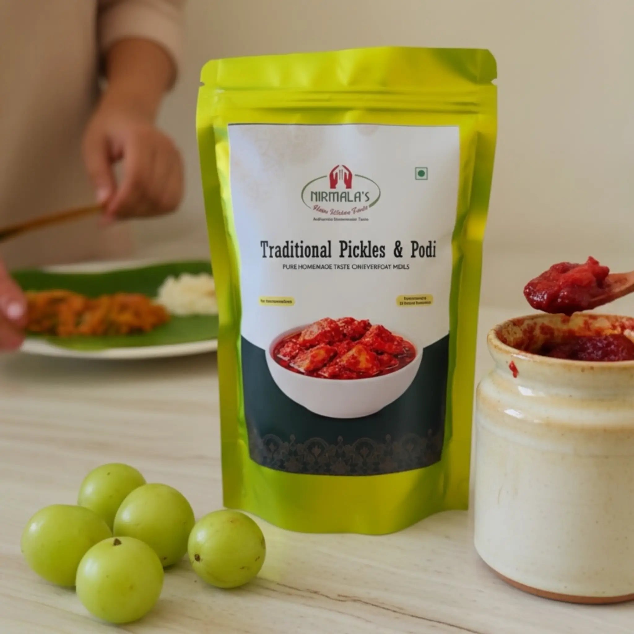 Green packet of Nirmala's Traditional Pickles & Podi with green amlas and a jar of pickle on a wooden surface.