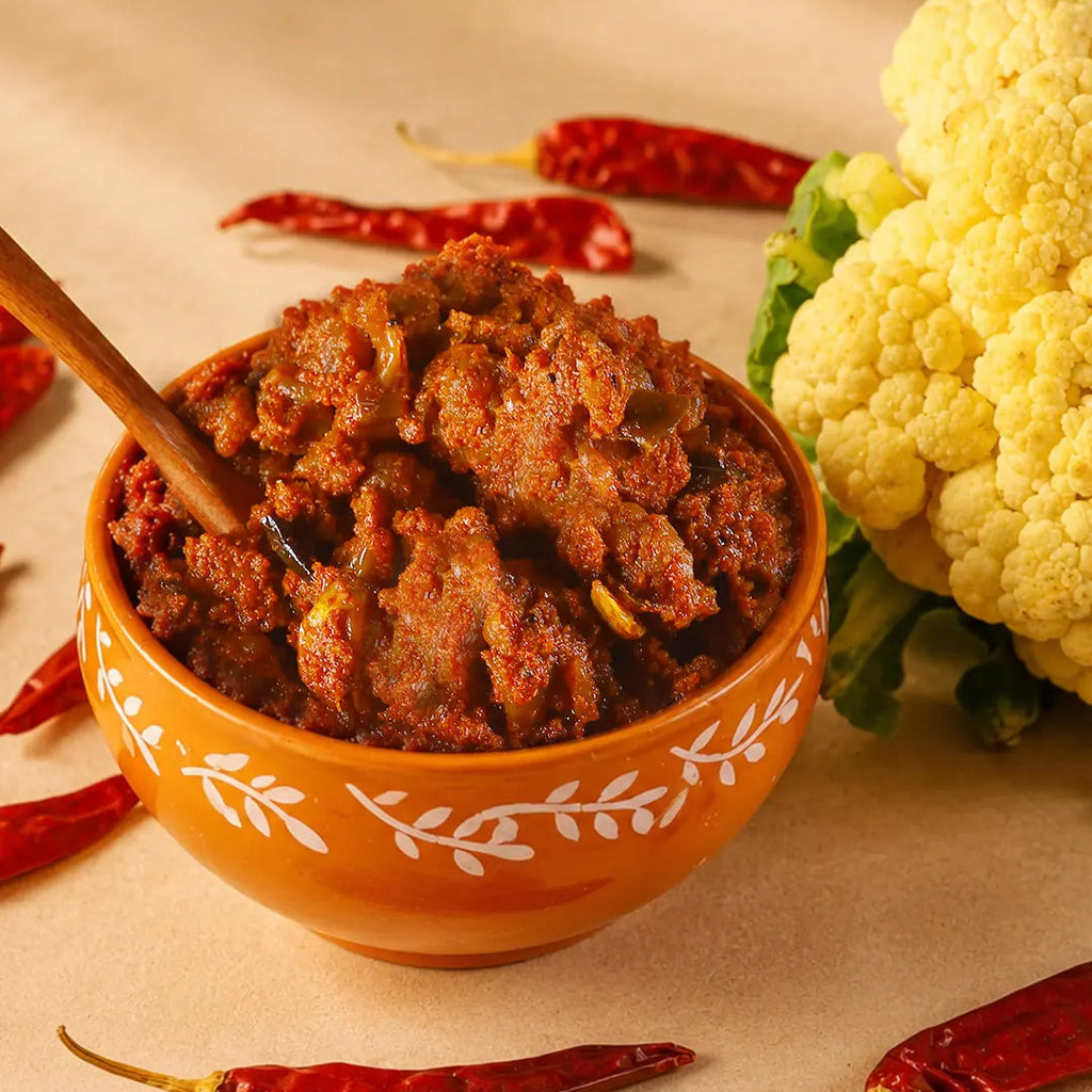 A close-up of a textured red-orange cauliflower pickle in an orange ceramic bowl with white leaf patterns, set on a beige surface with scattered red chilies and a yellow cauliflower nearby.