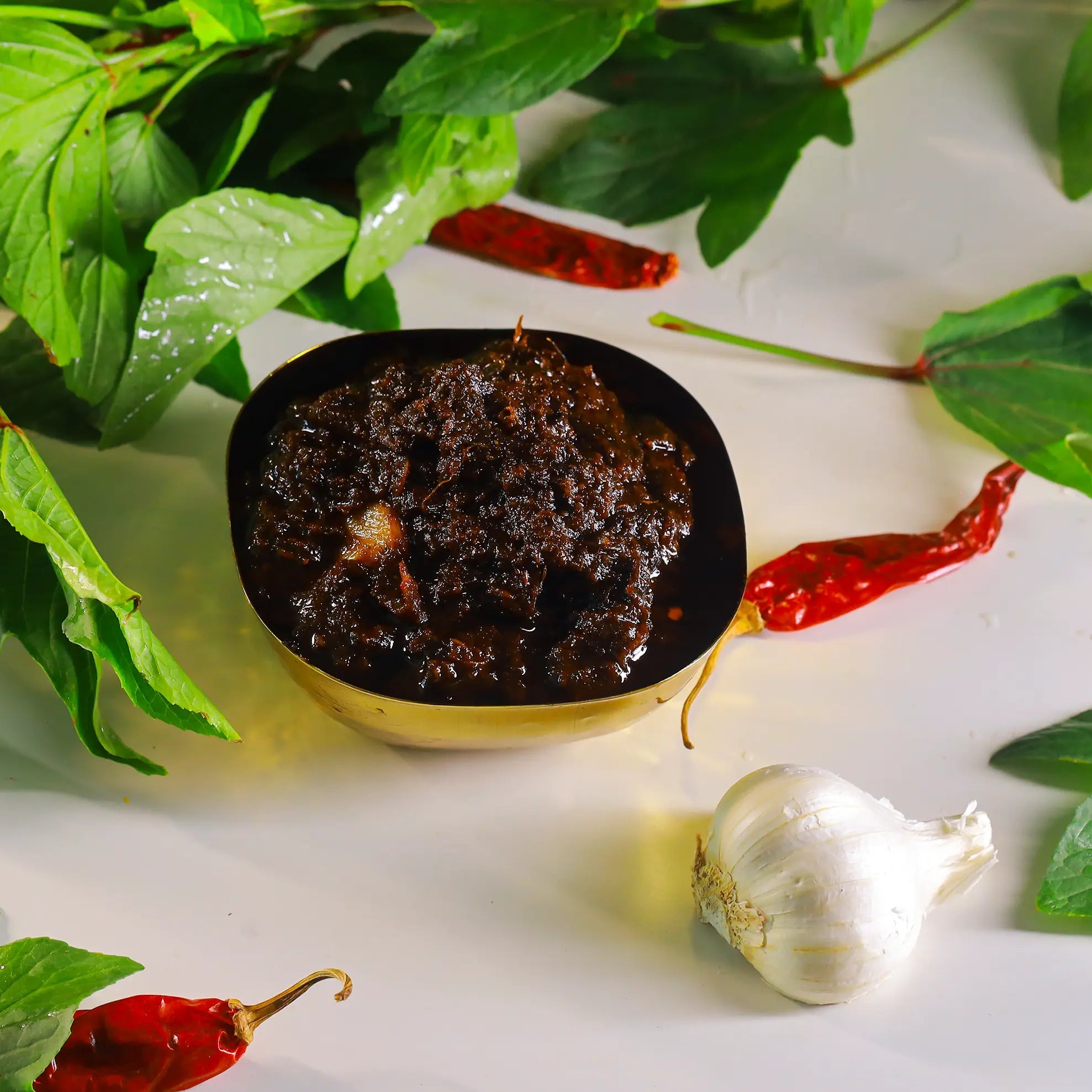A small bowl filled with a dark reddish-brown gongura pickle, styled with green gongura leaves, red chili peppers, and a garlic bulb on a white surface.
