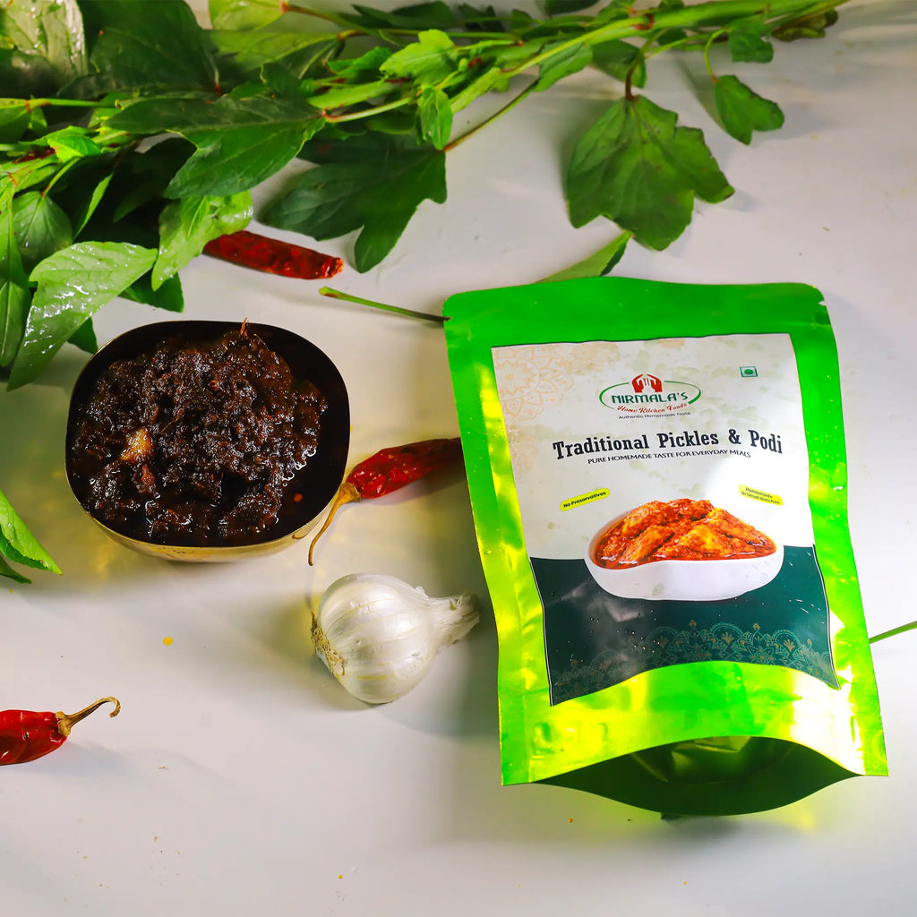 A green stand-up pouch of Nirmala's Traditional Pickles & Podi placed on a white surface with a bowl of gongura pickle, a garlic bulb, red chilies, and green herbs around it.