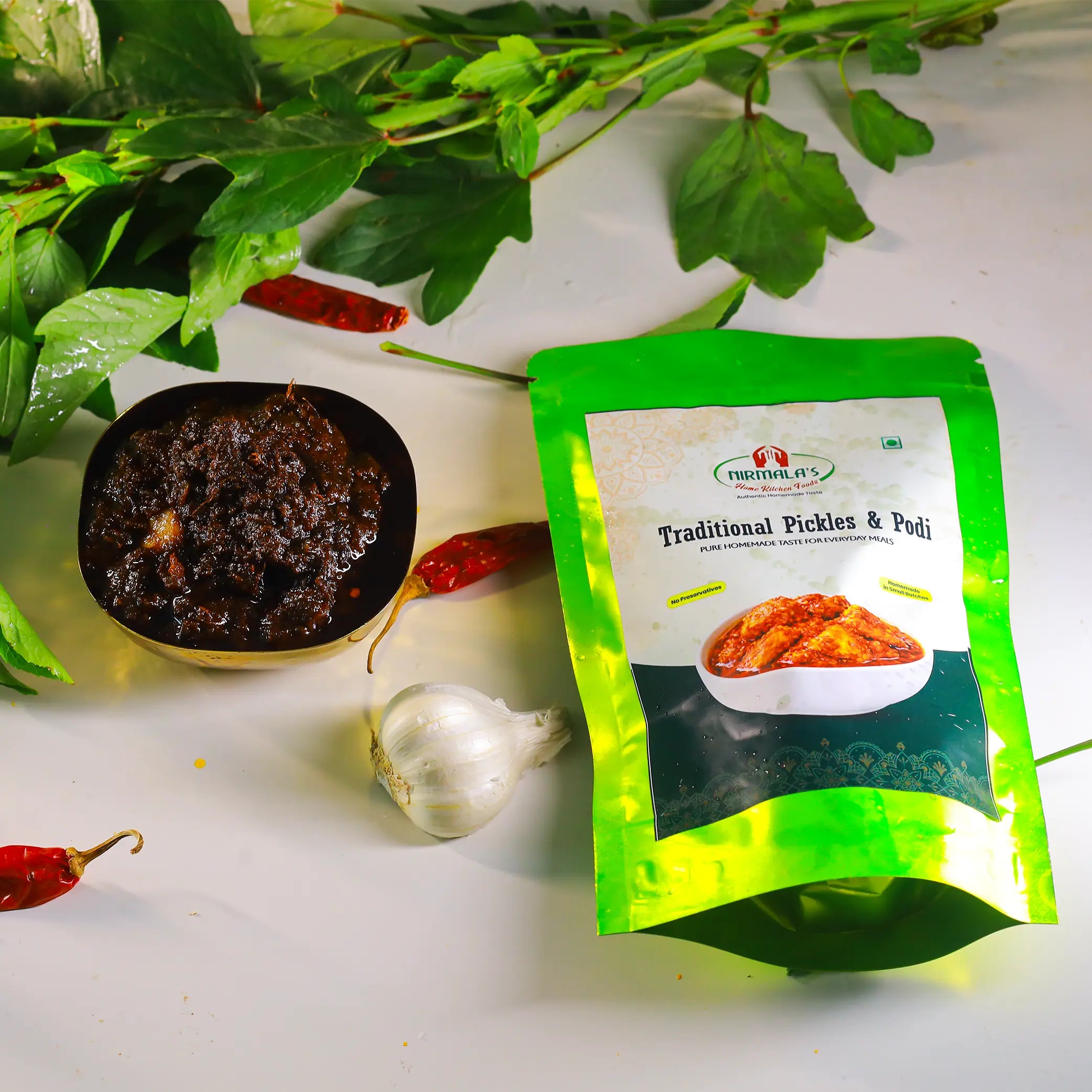 A green stand-up pouch of Nirmala's Traditional Pickles & Podi placed on a white surface with a bowl of gongura pickle, a garlic bulb, red chilies, and green herbs around it.