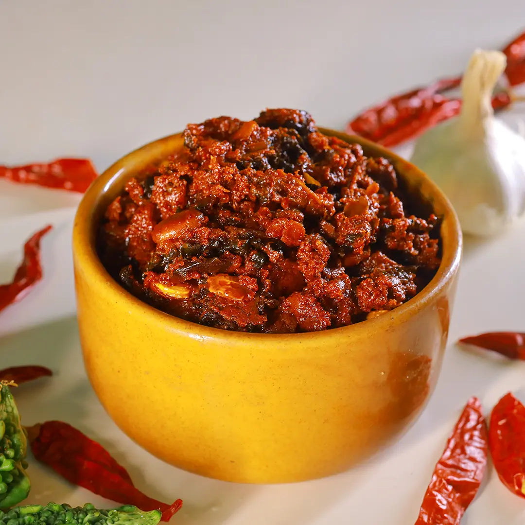 A yellow ceramic bowl filled with a chunky red-orange bitter gourd pickle, styled on a white surface with dried red chilies, a garlic bulb, and green vegetables around it.