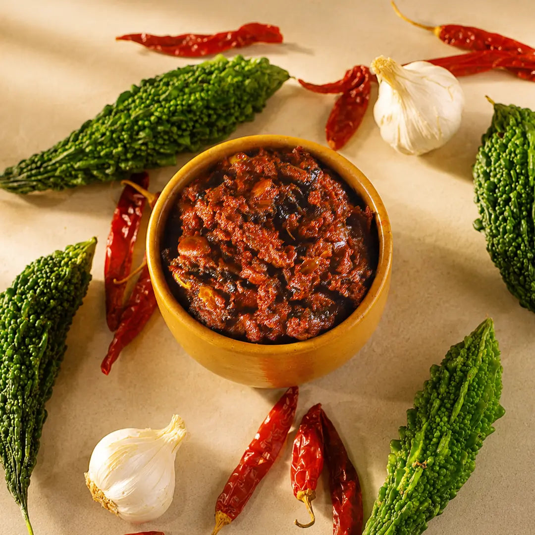 A small wooden bowl filled with a chunky red-orange kakarakaya pachadi, surrounded by green bitter gourds, red chilies and a garlic bulb on a beige surface.