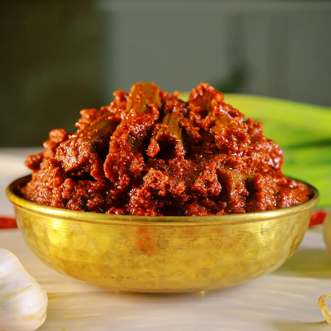 A close-up shot of a mound of reddish-brown textured drumstick pickle served in a shiny gold bowl with garlic and green leaves blurred in the background.