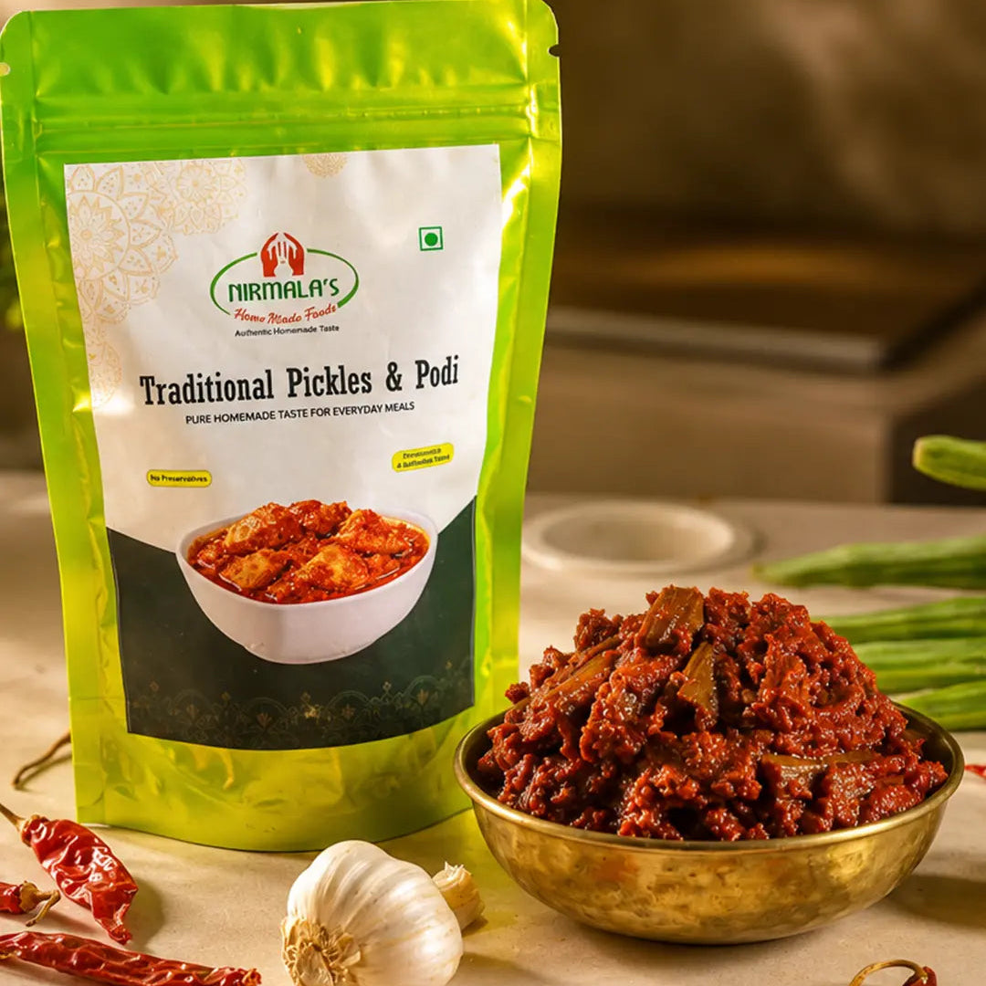 A stand-up pouch of Nirmali's Traditional Pickles & Podi in a bright green and white package, displayed next to a brass bowl filled with the reddish-brown Mulakkada pickle, with garlic, red chilies, and drumsticks arranged around it on a beige surface.