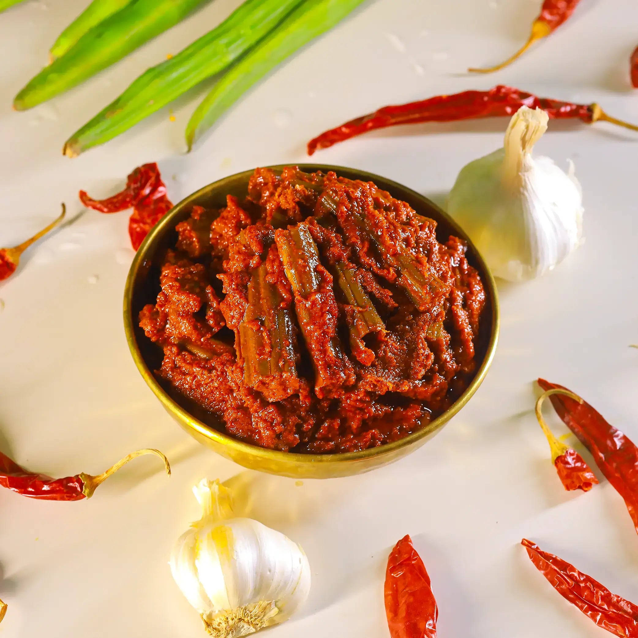 A small gold bowl filled with red cauliflower pickle, surrounded by whole garlic bulbs, dried red chilies, and green chili slices on a white surface.